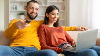 Mann und Frau sitzen auf dem Sofa, Frau hat Laptop auf dem Schoß, Mann hält Fernbedienung in der Hand und schaut TV. Beide lachen. 