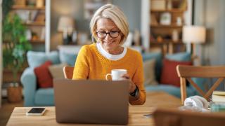 Eine blonde Frau mittleren Alters sitzt am Esstisch vor ihrem Laptop im Wohnzimmer. Sie hält eine weiße Tasse in ihrer Hand, schaut auf das Display und lächelt.