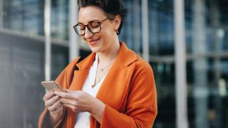 Eine Frau mittleren Alters mit Brille hält ihr Smartphone in den Händen, blickt darauf und lächelt mit offenem Mund. Sie trägt einen ockerfarbenen Mantel, hinter ihr ist ein verglastes Gebäude.