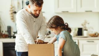 Ein Mann und Mädchen sind in der Küche, der Mann steht hinter der Küchentheke und das Mädchen sitzt darauf. Neben ihr steht ein Paket, das Vater und Tochter gemeinsam auspacken.